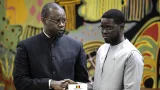 Diomaye Faye et Ousmane Sonko en tenue officielle se serrent la main devant un fond décoratif coloré