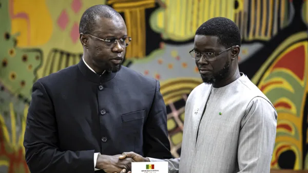 Diomaye Faye et Ousmane Sonko en tenue officielle se serrent la main devant un fond décoratif coloré