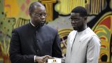 Diomaye Faye et Ousmane Sonko en tenue officielle se serrent la main devant un fond décoratif coloré